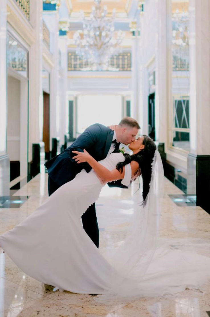 Groom dips his bride in a crepe fit and flare wedding dress with sleeves and a veil, and they kiss in a grand ballroom.