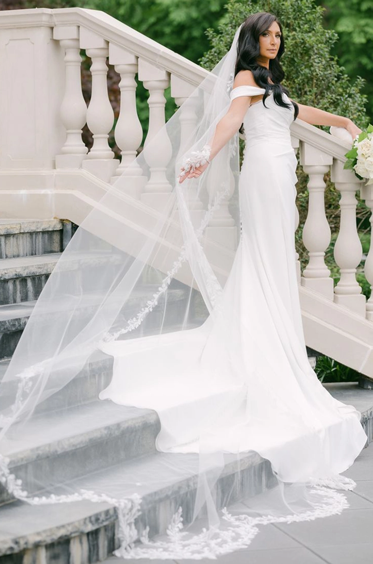 Bride poses on a staircase in a draped crepe bodice gown with sleeves and a lace-edge veil, holding a bouquet.