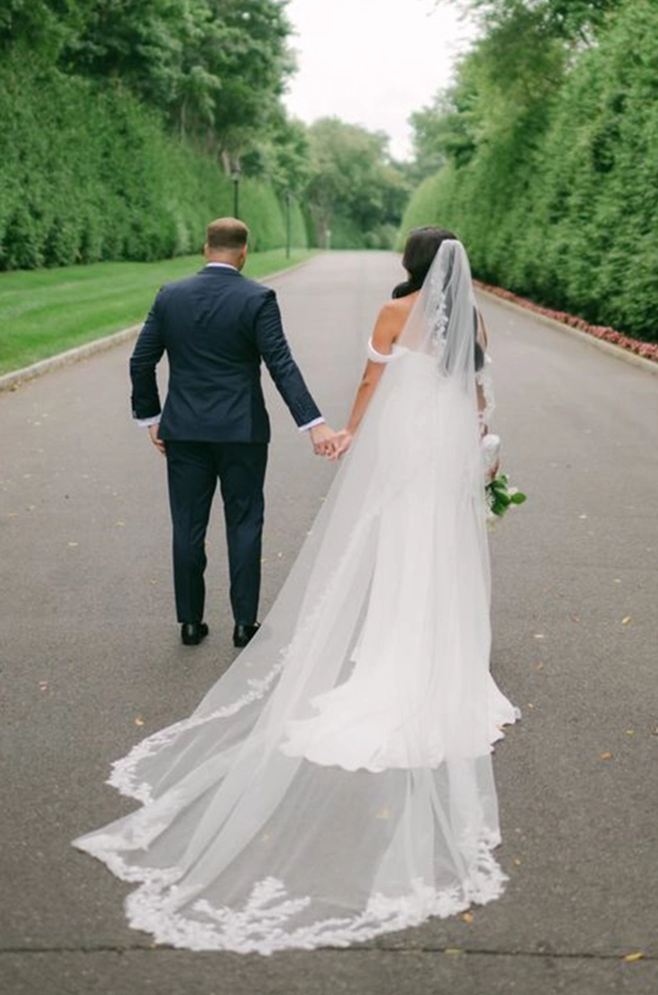 Back of bride in a crepe fitted gown with draped sleeves and a lace-edge veil, walks alongside her groom on a tree-lined path.