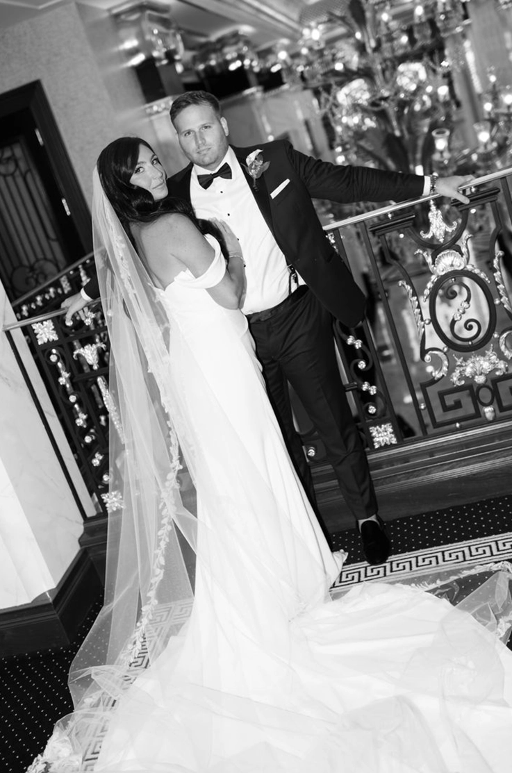 Bride in a crepe draped gown with sleeves and a lace-edge veil, poses with her groom leaning on a railing in black and white.