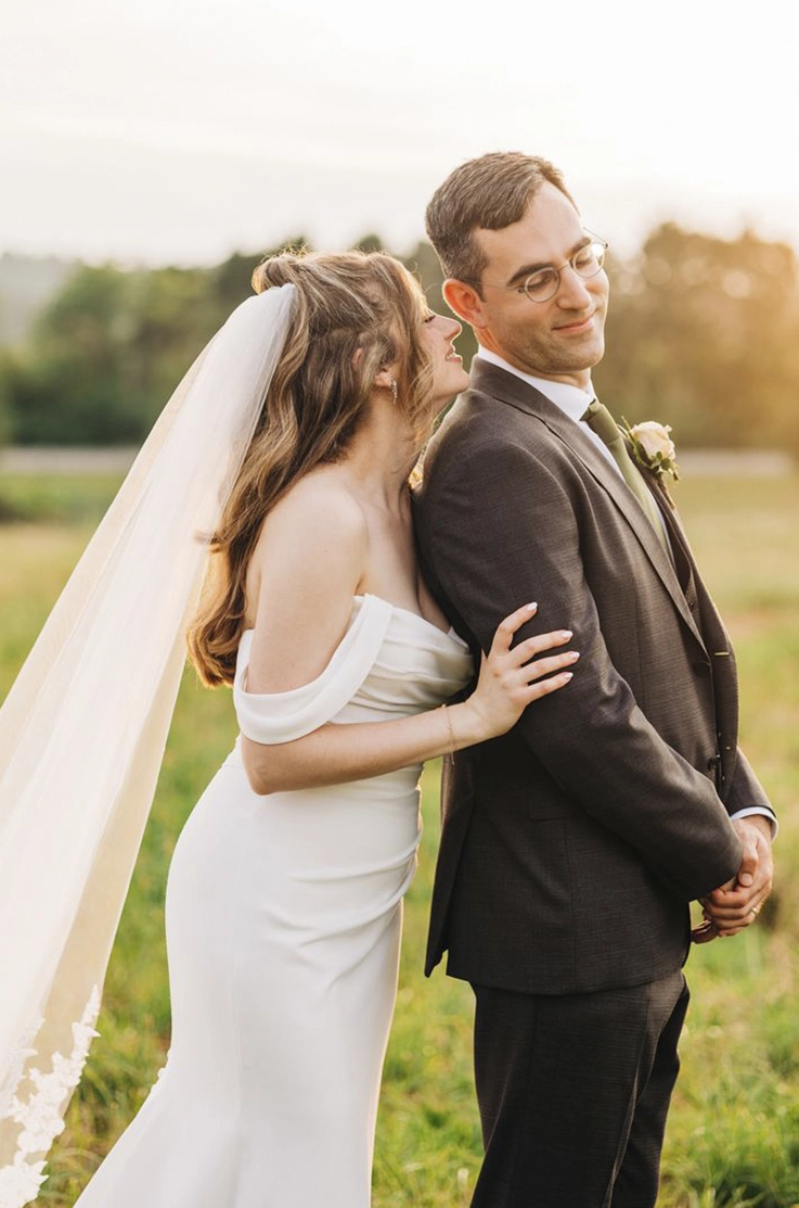 Close-up of bride in a draped crepe bodice gown with sleeves and a veil, embracing groom in a sunny open field. Close-up of bride in a draped crepe bodice gown with sleeves and a veil, embracing groom in a sunny open field. 