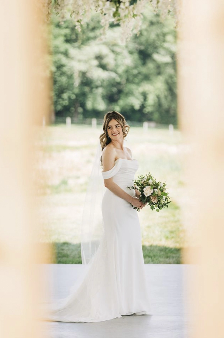 Bride poses in a crepe fitted gown with draping, sleeves and a veil, is holding a bouquet outside the venue.