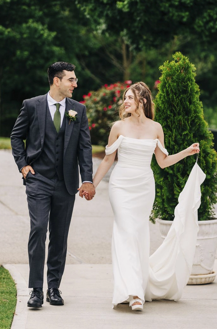 Bride in a fit and flare crepe wedding dress with a draped scoop neck, holds up her train while walking next to groom outdoors. Bride in a fit and flare crepe wedding dress with a draped scoop neck, holds up her train while walking next to groom outdoors.
