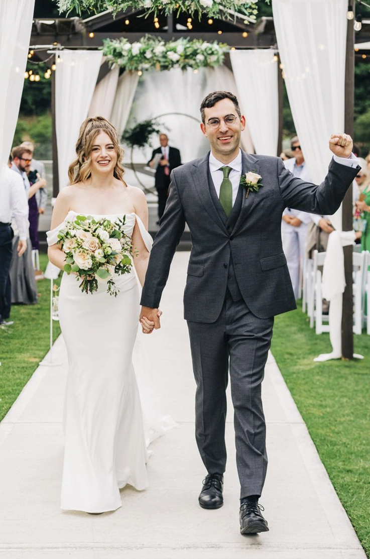 Bride in a fit and flare crepe gown with draped sleeves, holds a bouquet and walks down the aisle with her groom.