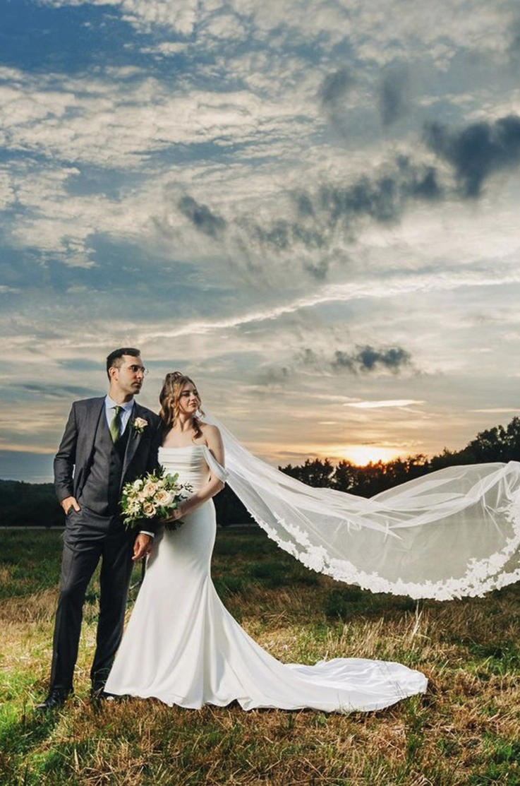Bride in a draped bodice gown with fit and flare skirt and a lace-edge veil, holds a bouquet beside the groom in a field. 