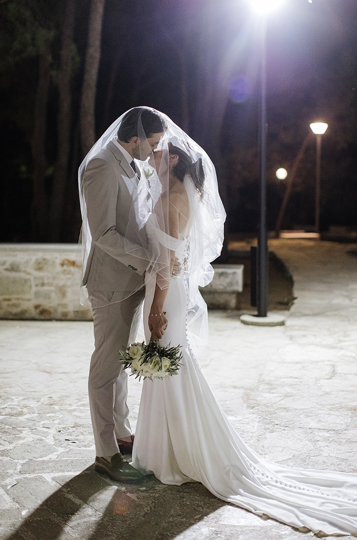 Bride in a fit and flare crepe gown with sleeves and buttons down the back, kisses groom under her veil at night.