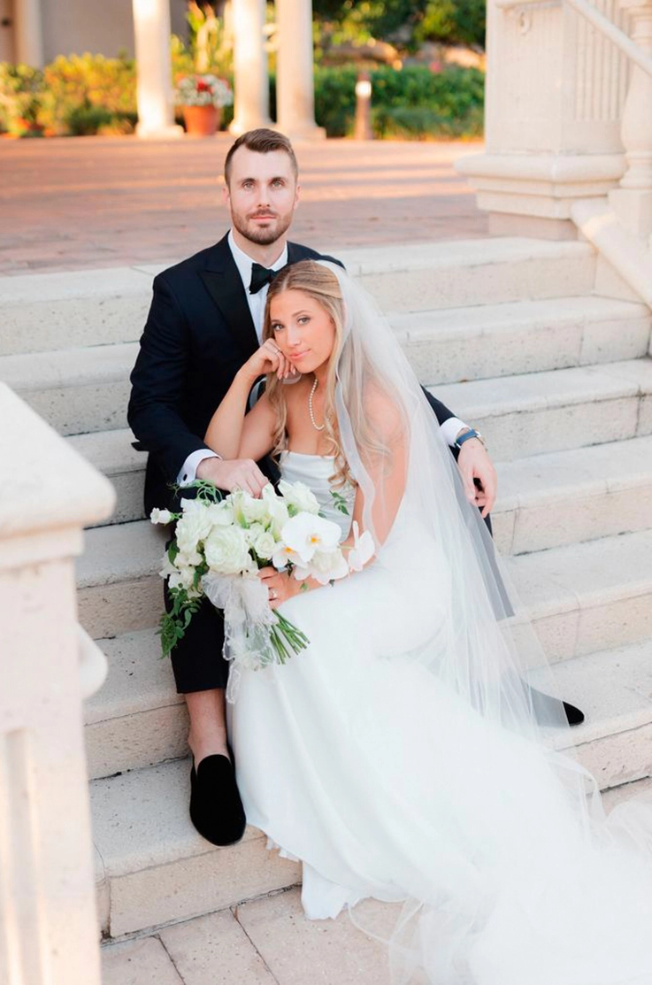 Bride in a strapless crepe gown with a draped scoop neckline and a veil, holds a bouquet in front of groom on stone steps.