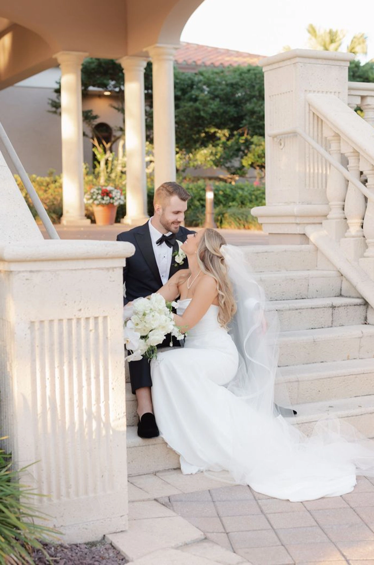 Bride in a sleek strapless wedding gown with a long flowing veil looks at her groom as they are sitting on outdoor steps.