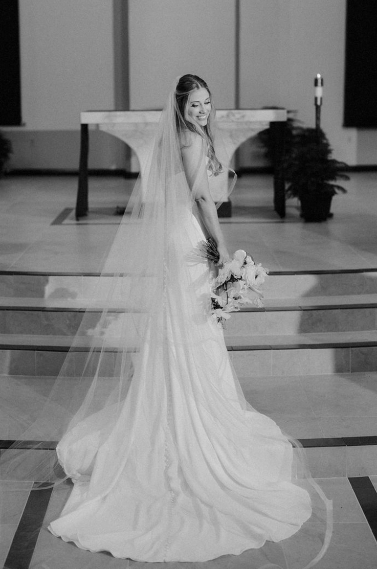 Bride poses in a crepe fit and flare gown with a flowing veil, holding a bouquet at a church altar, in black and white. 
