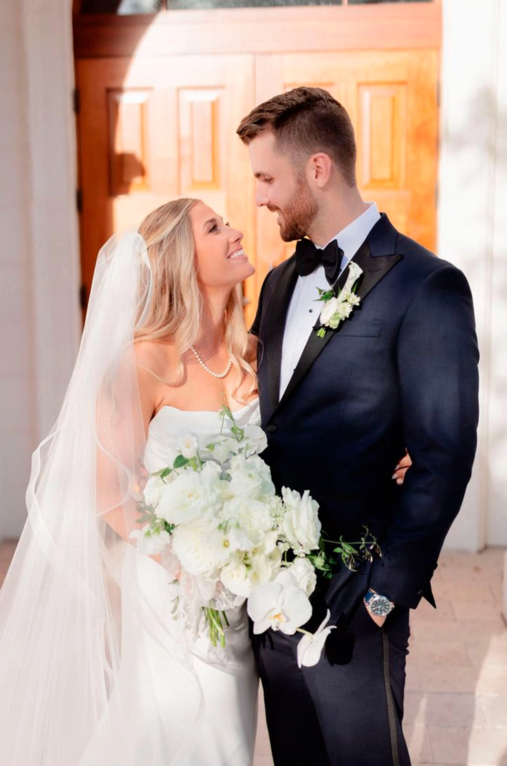 Bride in a draped crepe bodice gown with a scoop neck and veil, is holding a bouquet and faces groom in front of the venue.