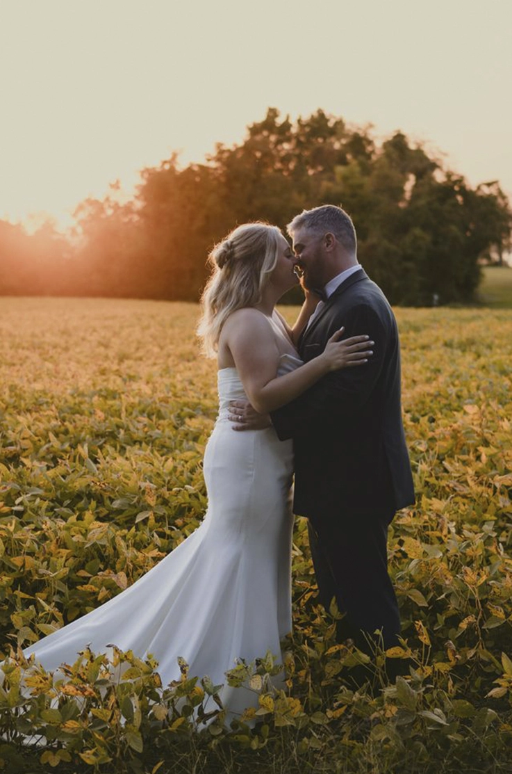 Bride in a strapless draped crepe bodice gown with a fit and flare skirt, faces groom in an embrace in a rural field.