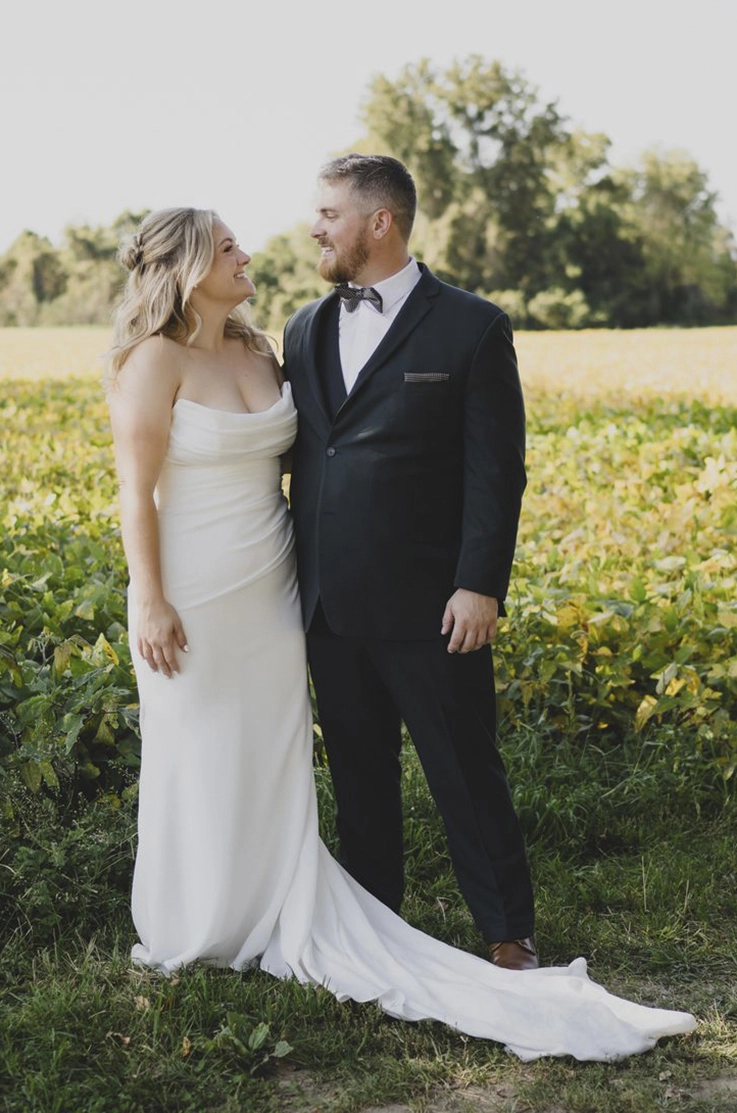 Bride in a strapless fit and flare gown with a draped scoop neckline, poses with her groom in a farm field