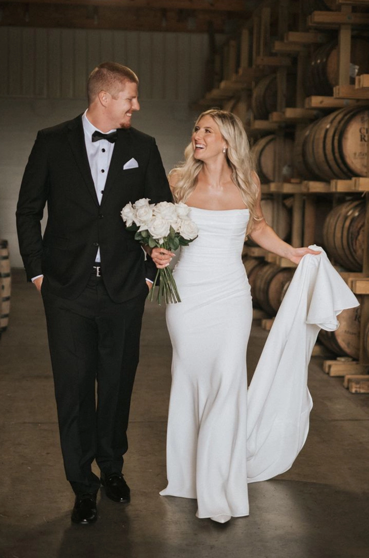 Bride in a fit and flare crepe gown with a draped scoop neck, picks up her train and walks with groom at a barrel wall.