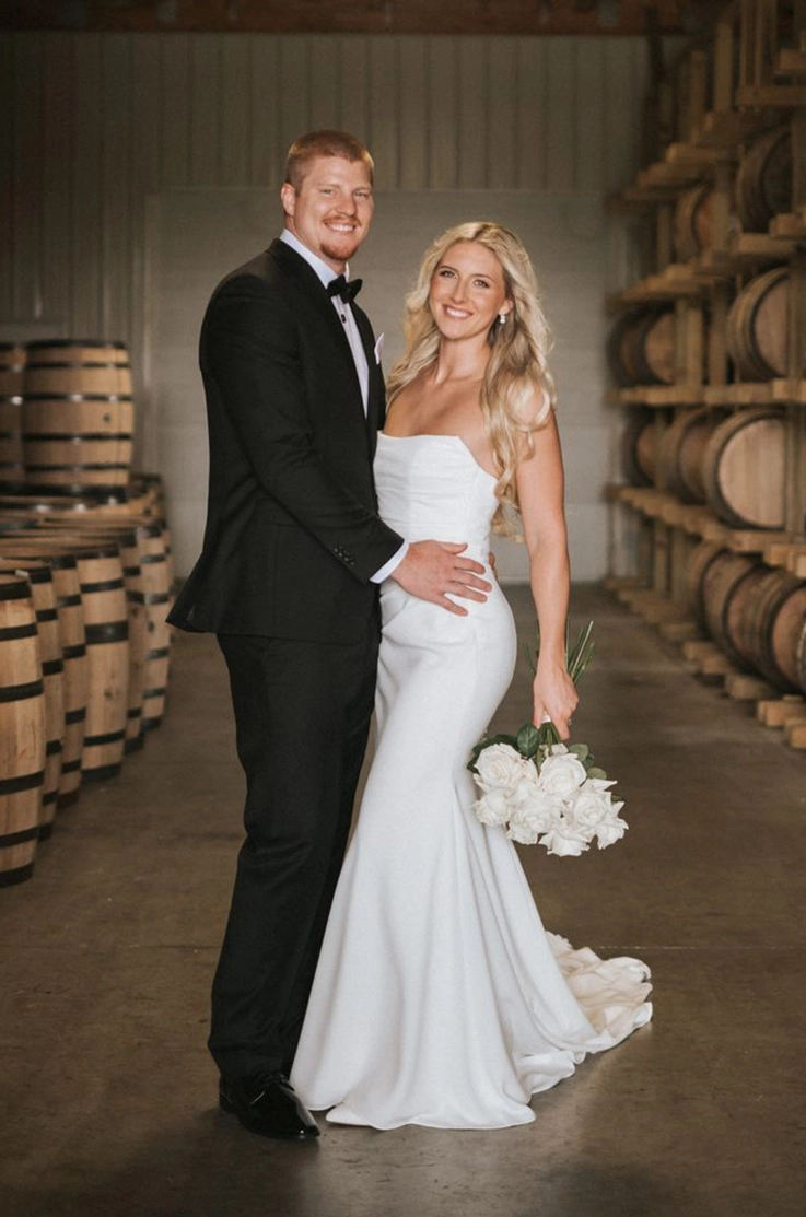 Groom poses with bride in a fit and flare crepe gown with a draped scoop neck, holding a white rose bouquet by a barrel wall. 