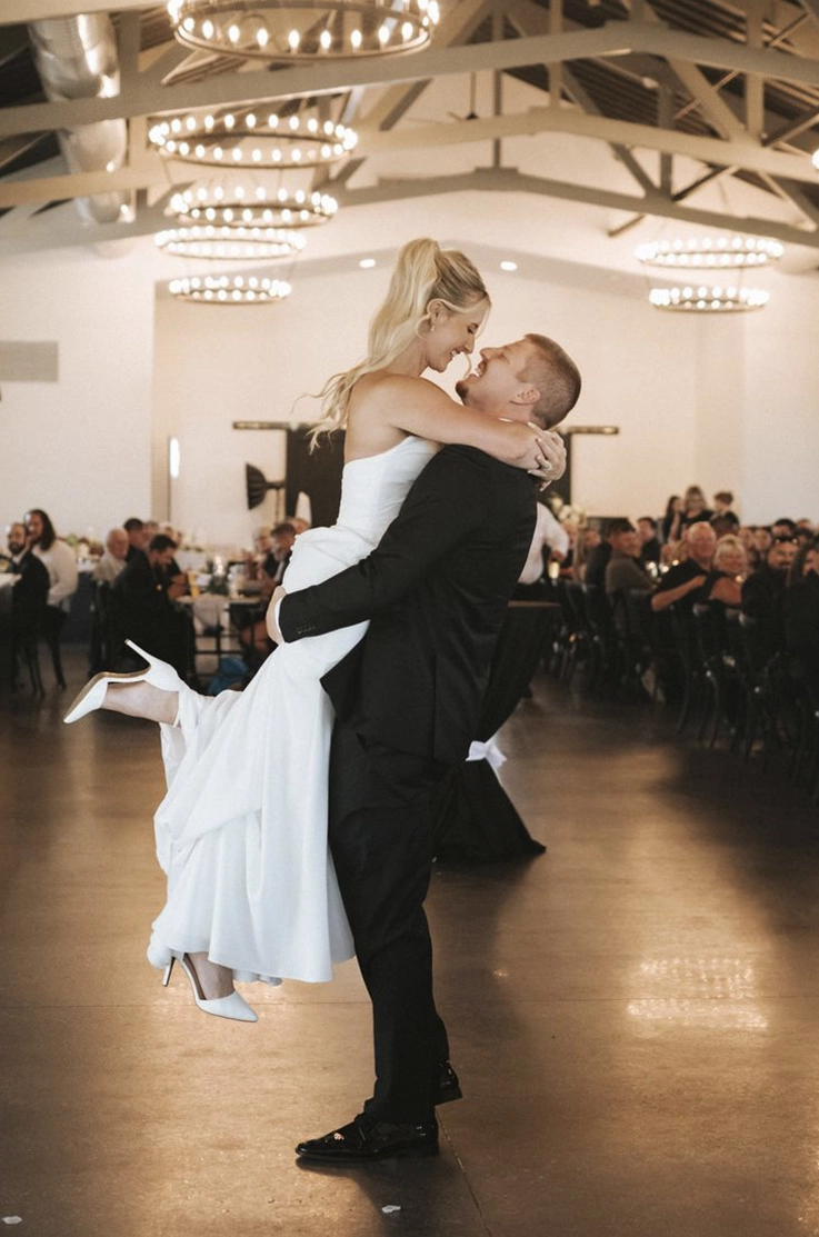 Groom picks up his bride in a strapless crepe wedding dress, in front of guests in a vaulted indoor reception hall.