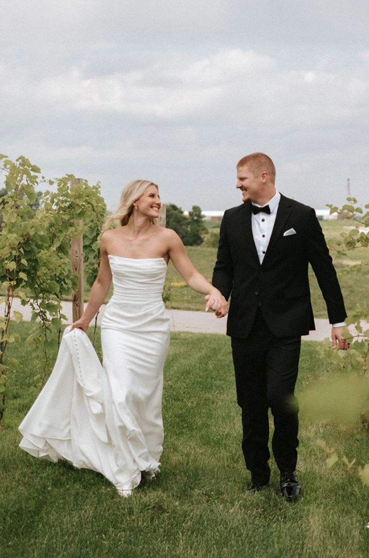 Bride in a crepe draped bodice gown with fit and flare skirt and buttons, walks hand in hand with her groom though a vineyard.