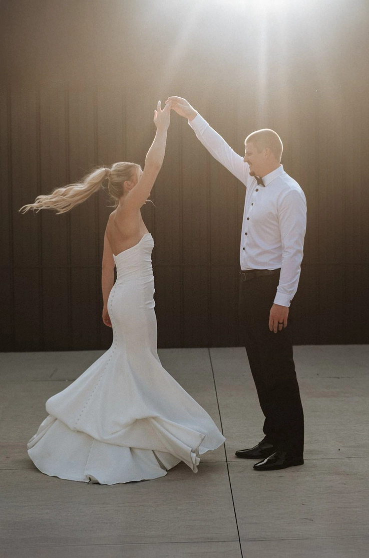 Groom twirls his bride in a strapless crepe fitted gown with a draped bodice and buttons, outside the venue in the sunlight.