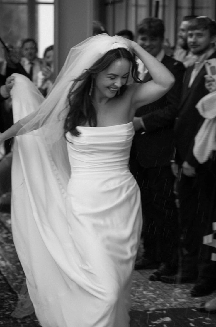Bride in a draped fit and flare wedding dress with a veil, joyfully celebrates among guests in black and white.