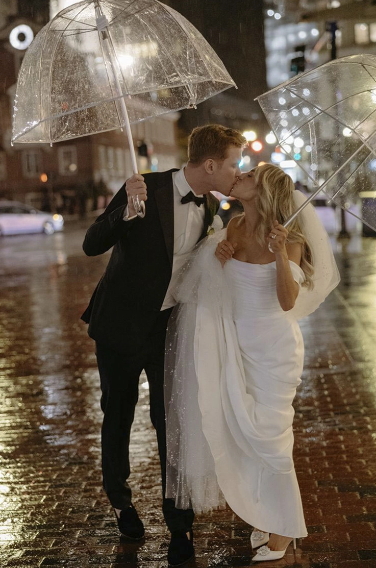 Bride in a crepe fit and flare gown with a draped bodice and sleeves, kisses groom under umbrellas on a rainy city street.