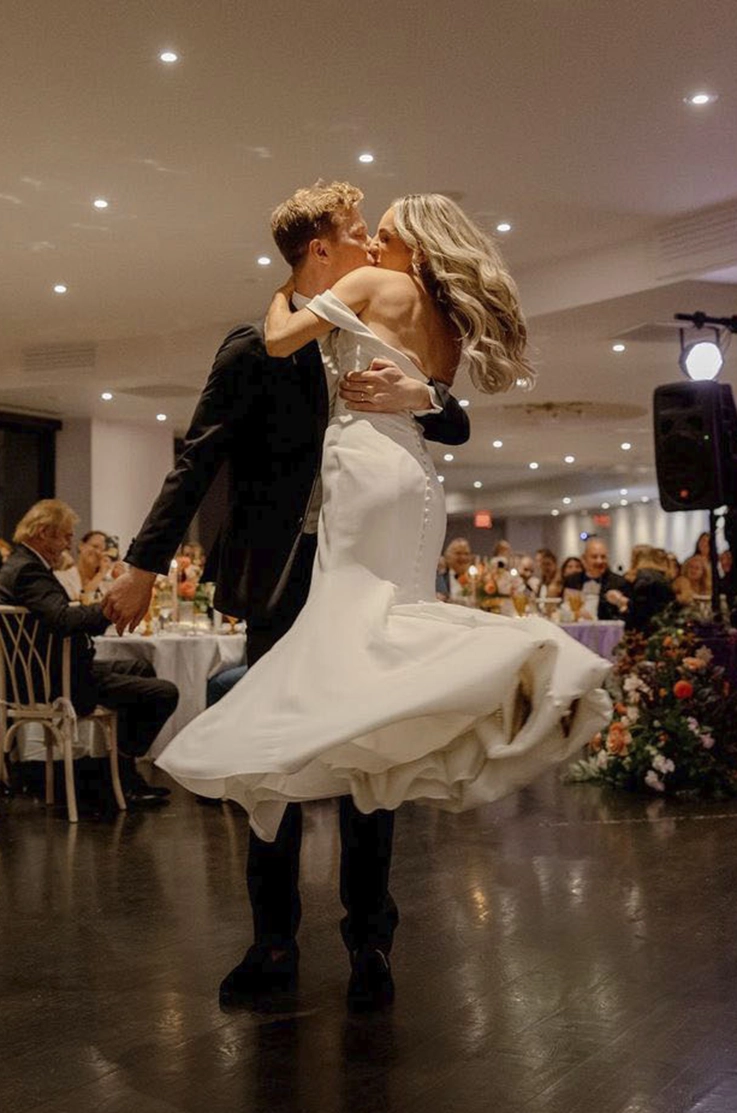 Groom picks up his bride in an off-shoulder crepe wedding dress with fit and flare skirt and sleeves, as they dance.