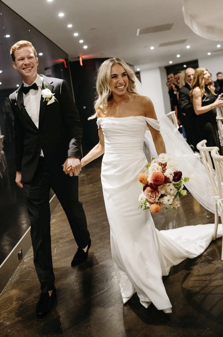 Bride in a crepe fitted gown with a scoop neck, draping and a veil, walks hand in hand with groom at a modern venue. 