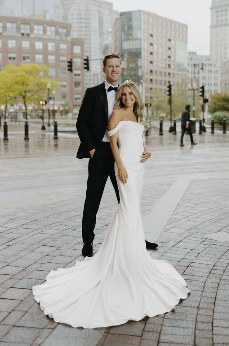 Bride poses in a draped crepe fitted gown with a draped bodice and sleeves, beside groom on a city street.
