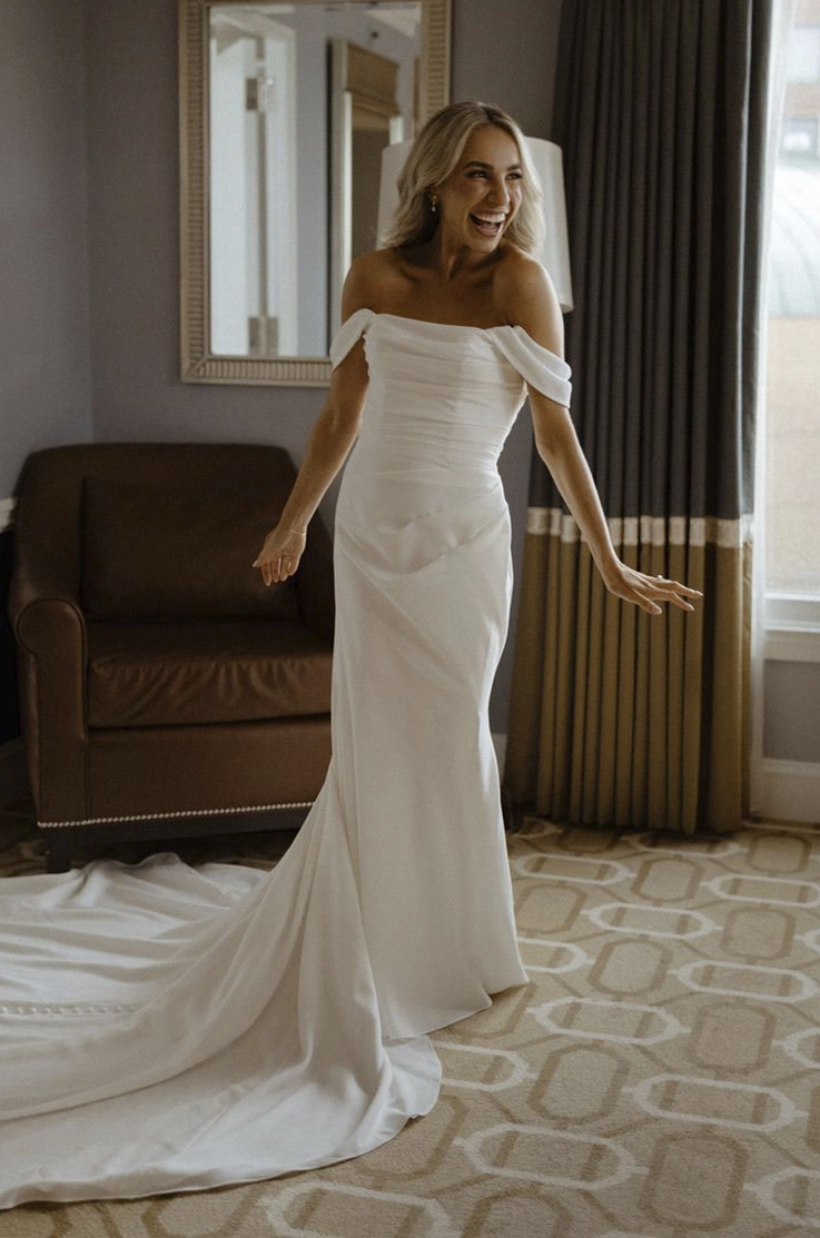 Bride in an off-shoulder draped bodice gown with fit and flare skirt and buttons, smiling in a bridal suite.