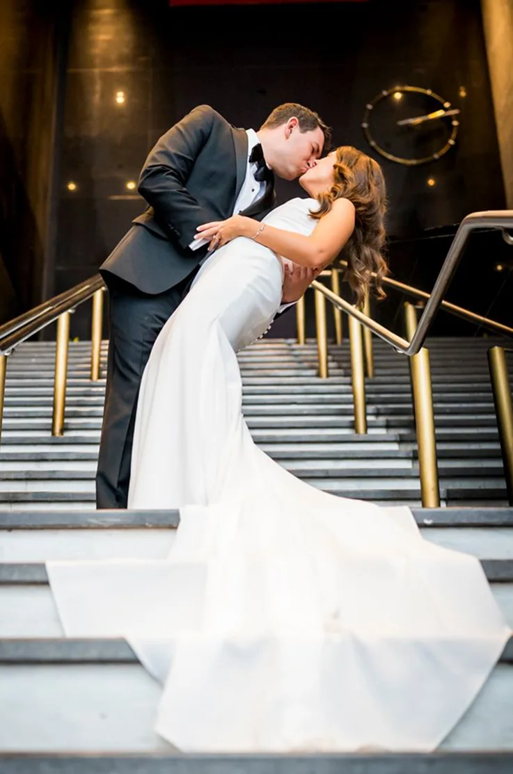 Groom dips his bride in a crepe fit and flare wedding dress on a grand staircase with gold railings. 