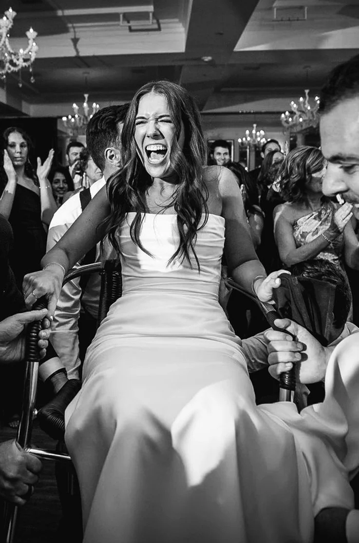 Bride is lifted up in a chair, in a strapless ruched gown with a fit and flare skirt and draping, shown in black and white.