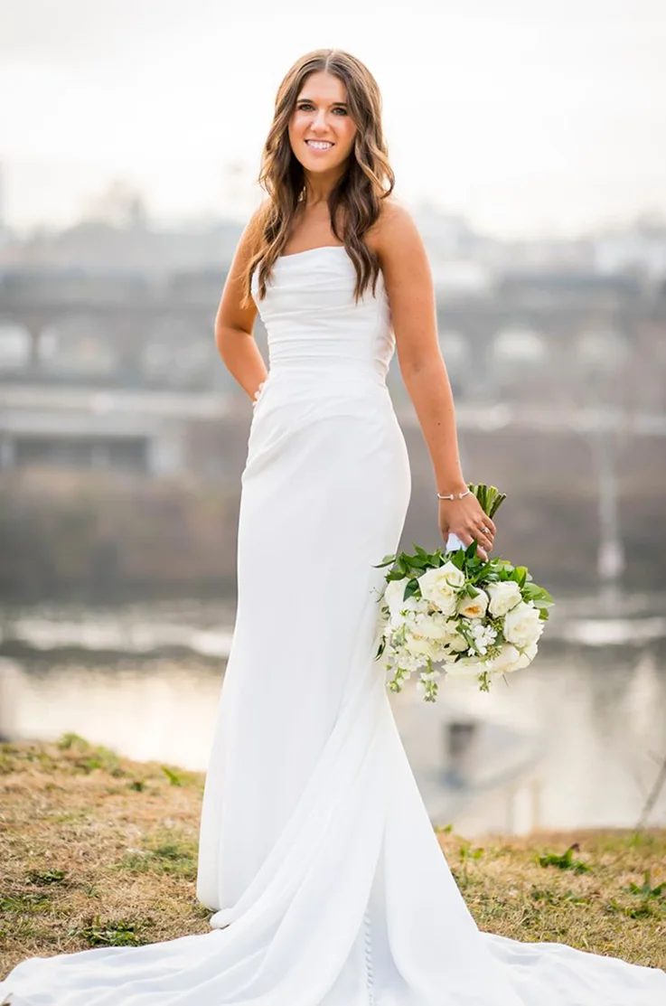 Bride poses in a crepe fit and flare gown with a draped bodice and a scoop neck, holding a bouquet, by a lake outdoors.