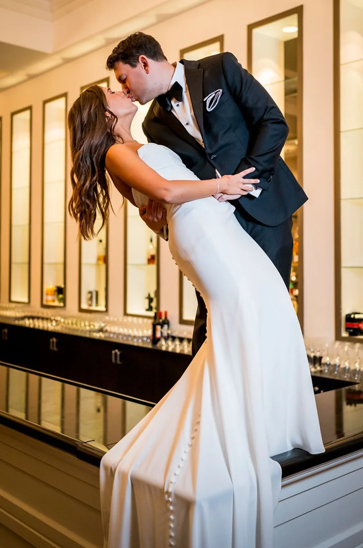 Groom dips his bride in a crepe fitted gown with buttons down the back, and they kiss on a bar countertop with warm lighting.