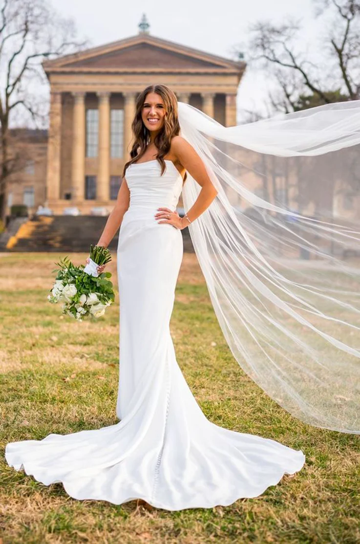Bride poses outside the venue in a crepe draped bodice gown with fit and flare skirt and a flowing veil, holding a bouquet
