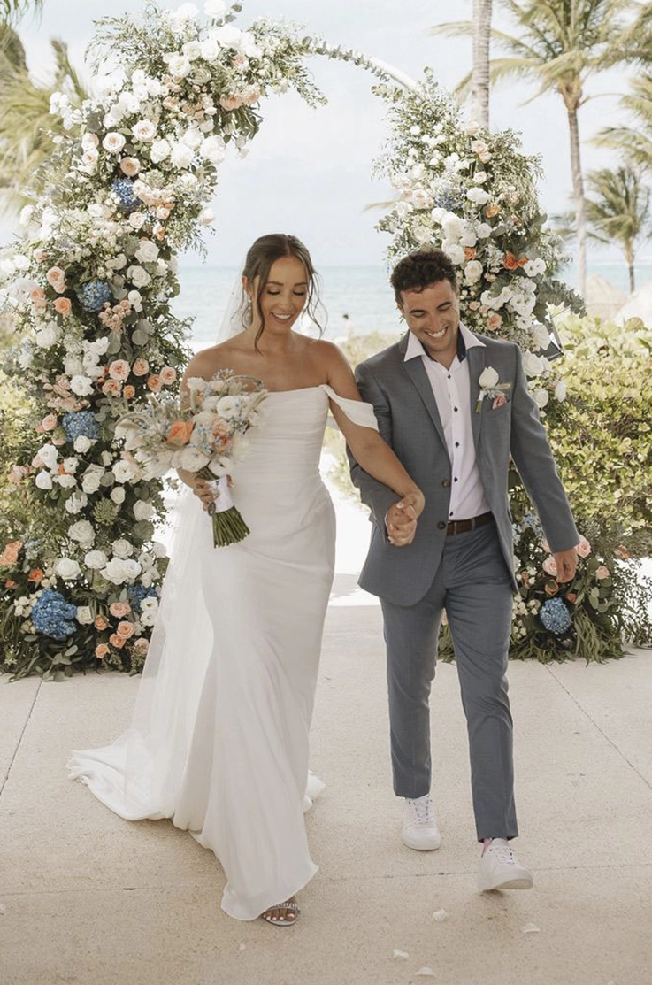 Bride in a draped crepe bodice gown with off-shoulder sleeves and a veil, walks alongside her groom under a floral arch.