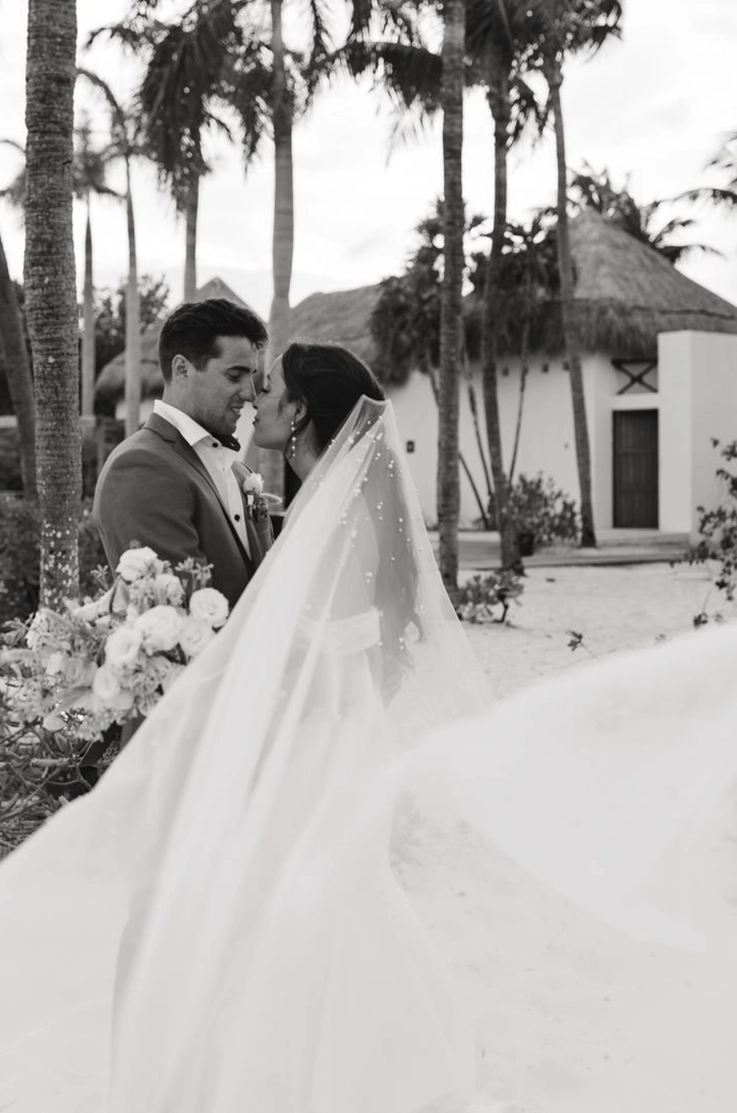 Groom faces bride in a crepe fit and flare gown with draped sleeves and a pearl veil, at a tropical resort in black and white.
