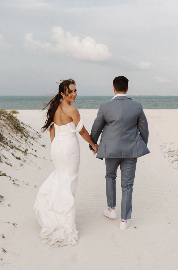 Bride in a crepe fitted gown with a draped bodice and sleeves, walks hand in hand with her groom on a white sand beach.