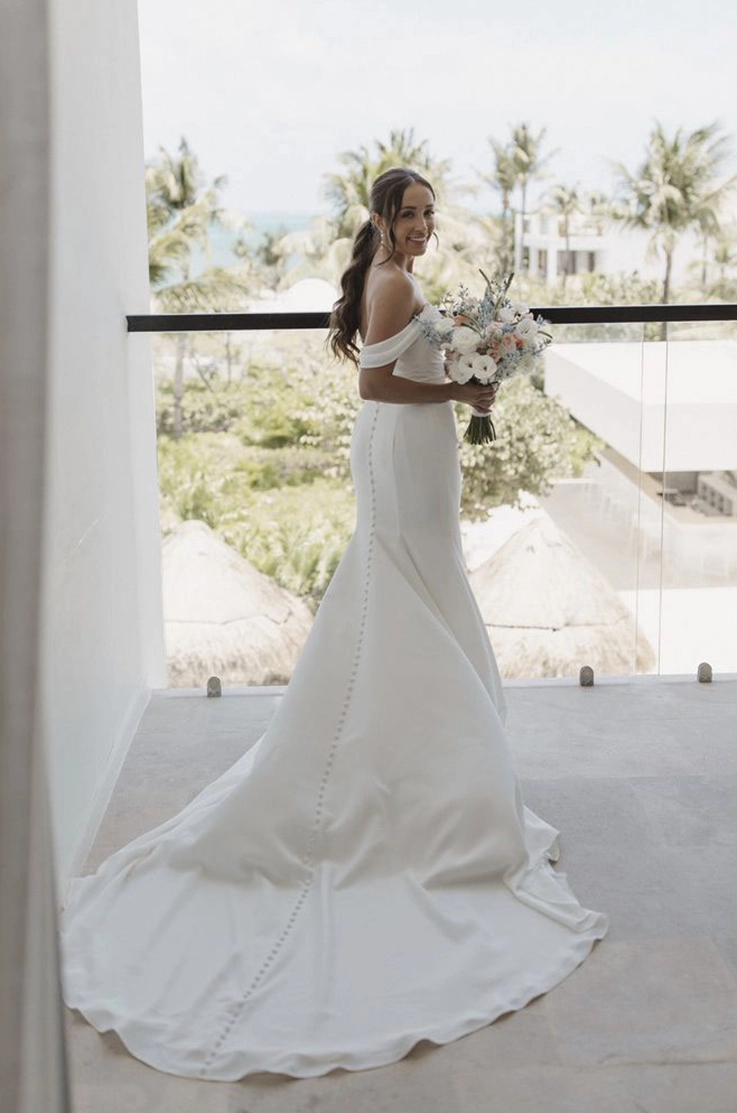 Bride poses in a fit and flare crepe gown with draped sleeves and buttons down the train, holding a bouquet on a balcony.
