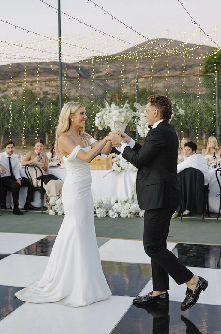 Bride in a scoop neck crepe wedding dress with draped sleeves, dances with her groom on a checkered dance floor under lights.