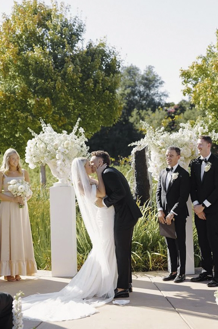 Groom kisses his bride at the altar, in a strapless fit and flare gown with draping, sleeves and a pearl veil.