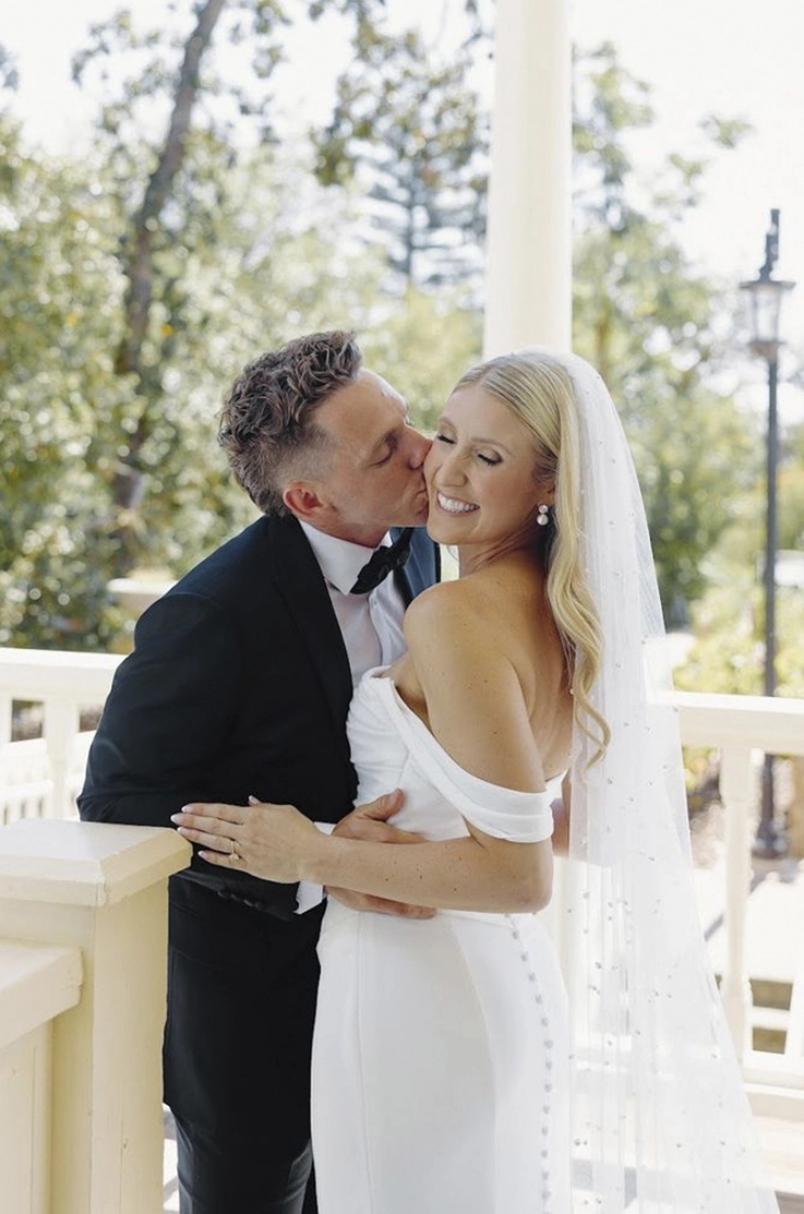 Groom holds his bride in an embrace, in a fitted crepe gown with a draped bodice, buttons and a veil by a white railing.