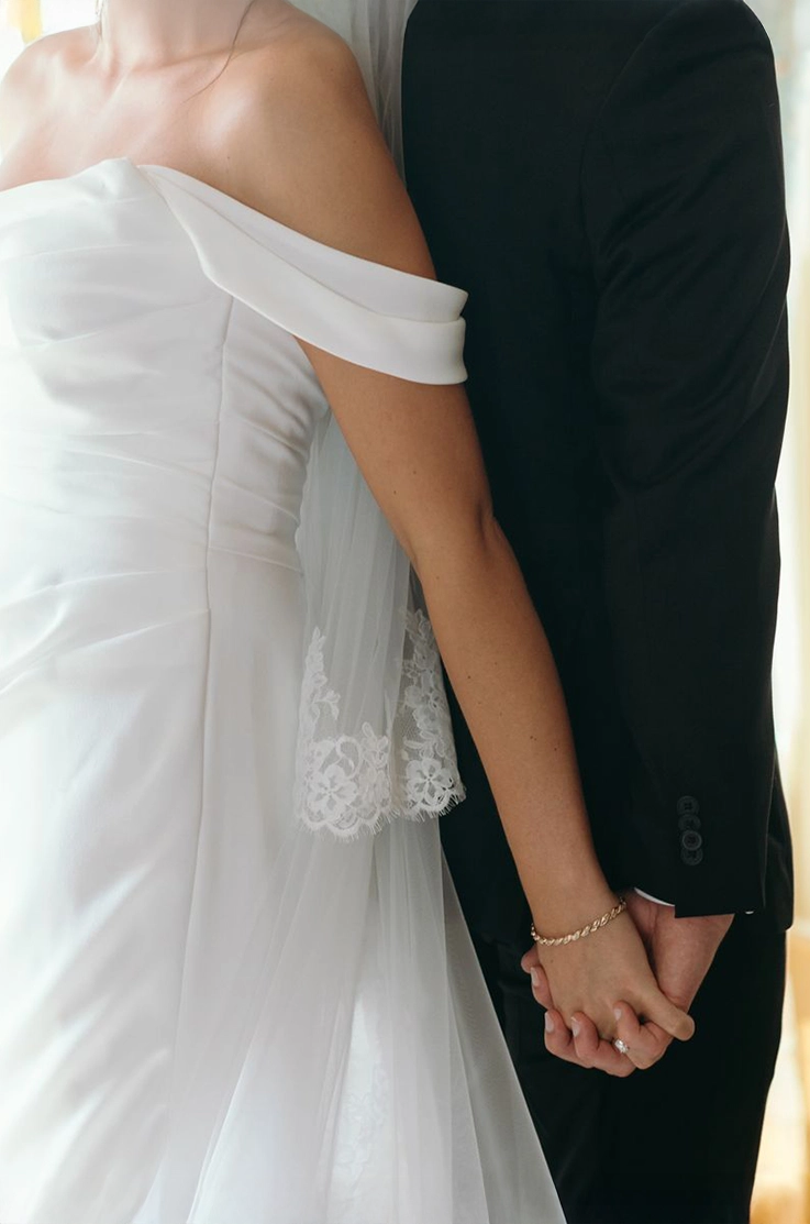 Close-up of bride in a draped crepe bodice gown with sleeves and a lace-trim veil, holding hands with the groom.