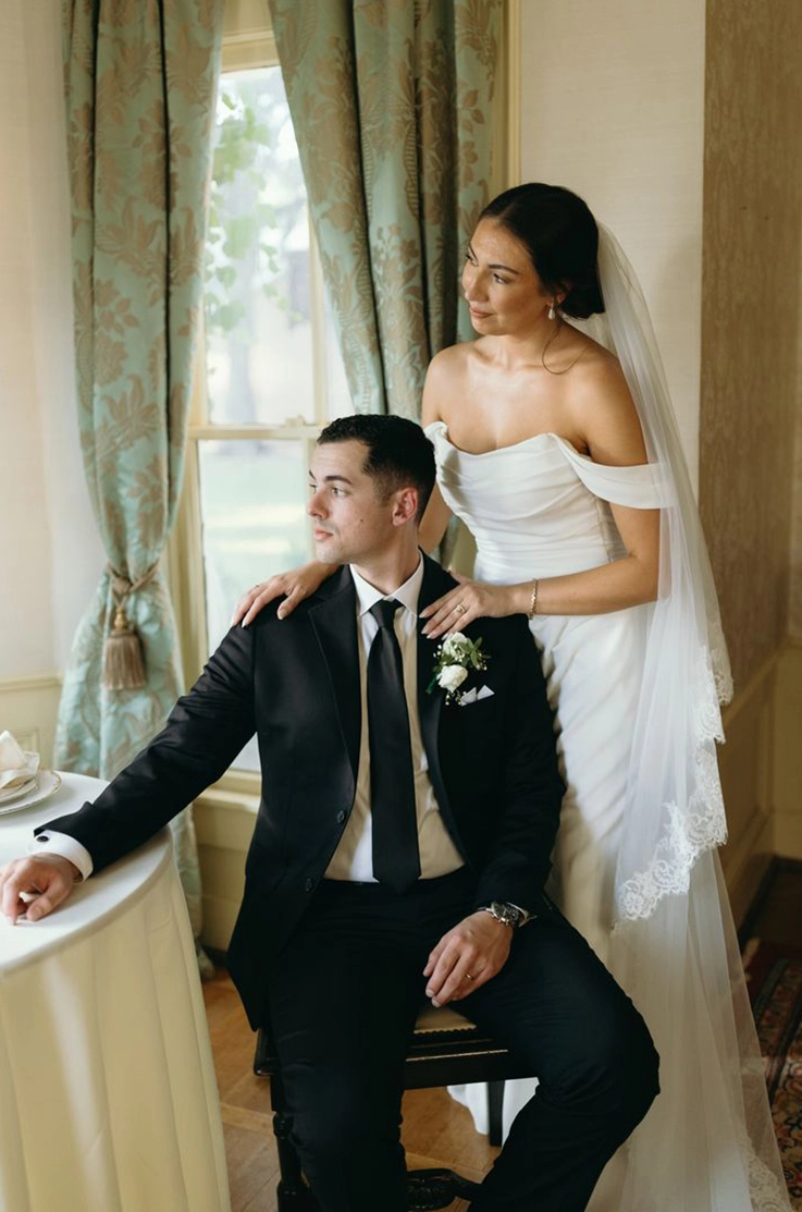 Bride in a crepe fitted gown with draping, sleeves and a veil poses with groom in a black suit sitting in a chair by a window.
