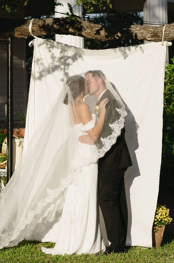 Groom kisses his bride in a fit and flare crepe gown with draped sleeves, under a lace-trim veil in front of a hanged curtain.