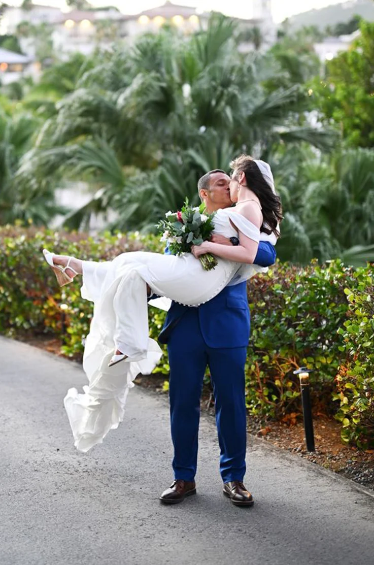 Groom picks up his bride in a crepe fitted gown with a buttoned back, draped sleeves and veil, and kiss on a path in a resort.