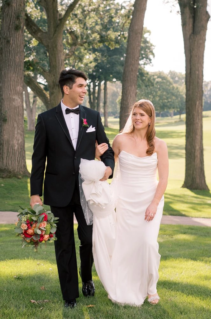 Groom walks alongside bride, holding her train in a crepe fitted gown with draping, between trees in an open sunny field. 