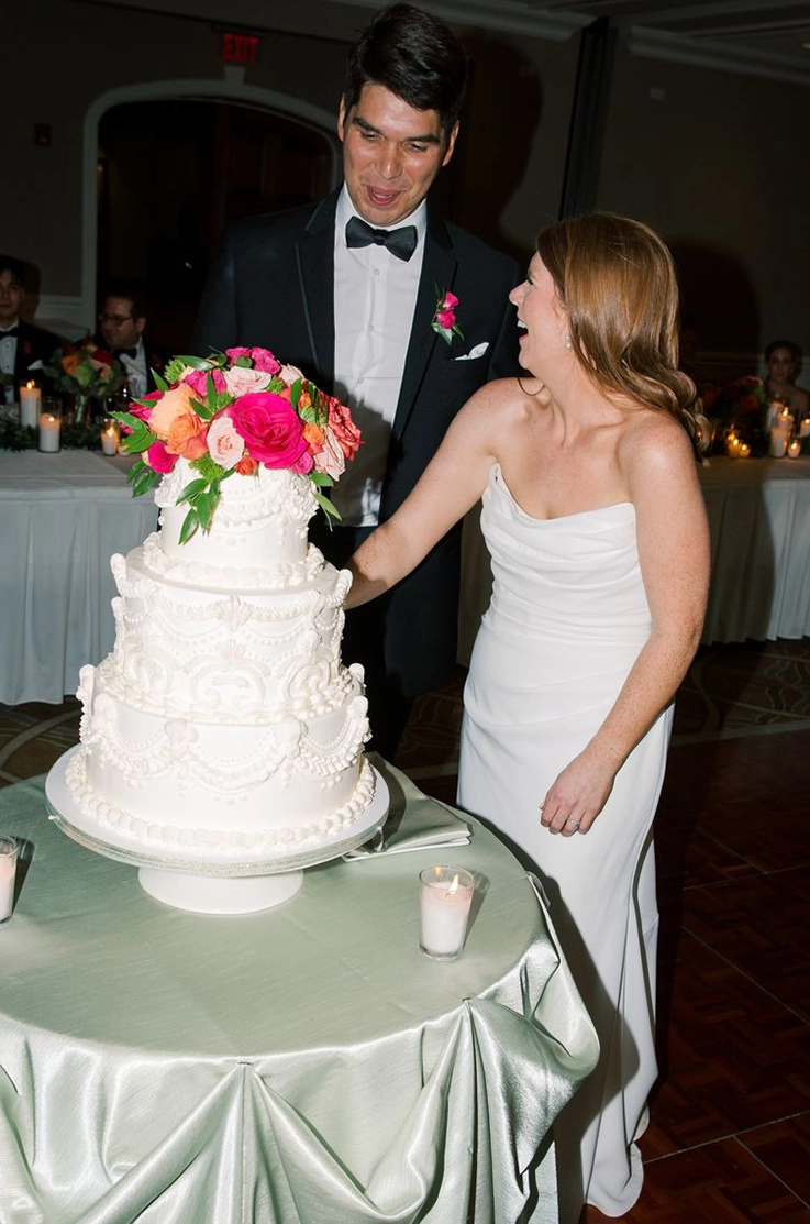 Bride in a draped crepe bodice gown with a scoop neck, cuts cake with her groom in a black tuxedo at candlelight reception.