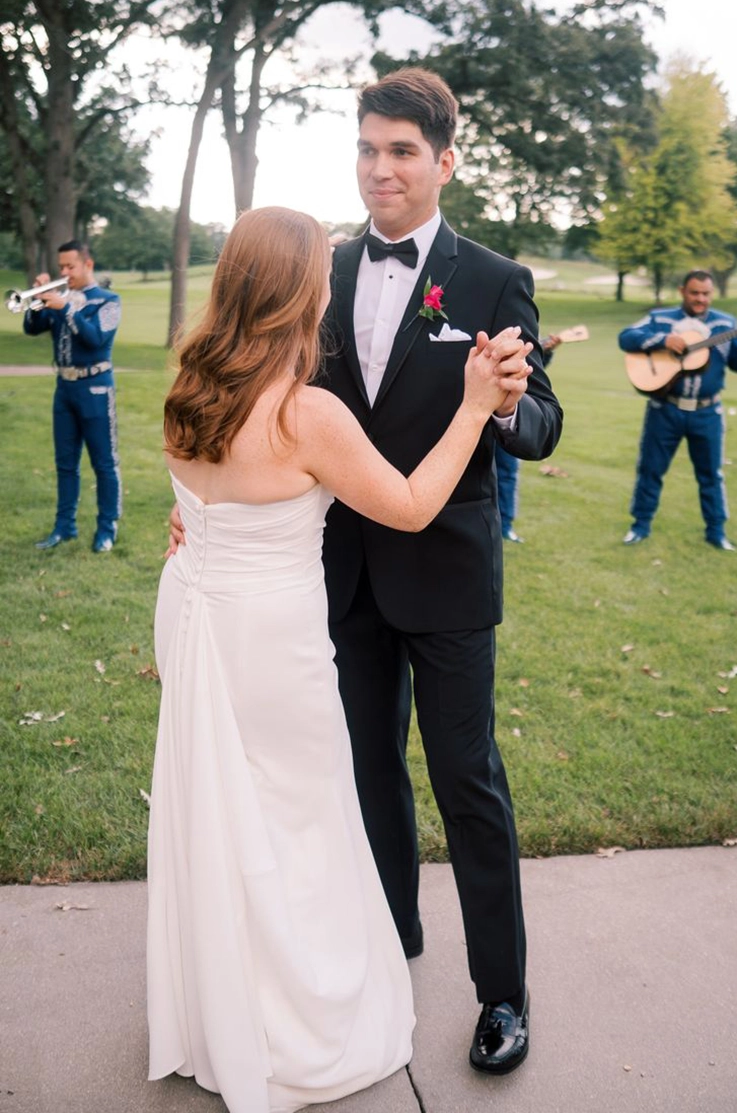 Groom dances with his bride in a strapless draped bodice gown with a buttoned back near a mariachi band outdoors.
