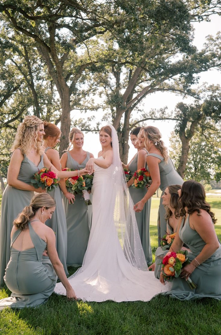 Bride in a fit and flare crepe gown with a draped scoop neck and a veil, poses in the middle of her bridesmaids holding bouquets.