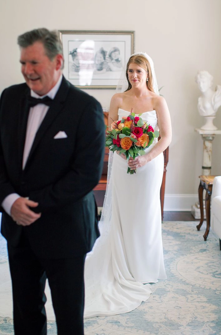 Bride in a strapless crepe fitted gown with a draped bodice and veil, holds a colorful bouquet indoors by her father.