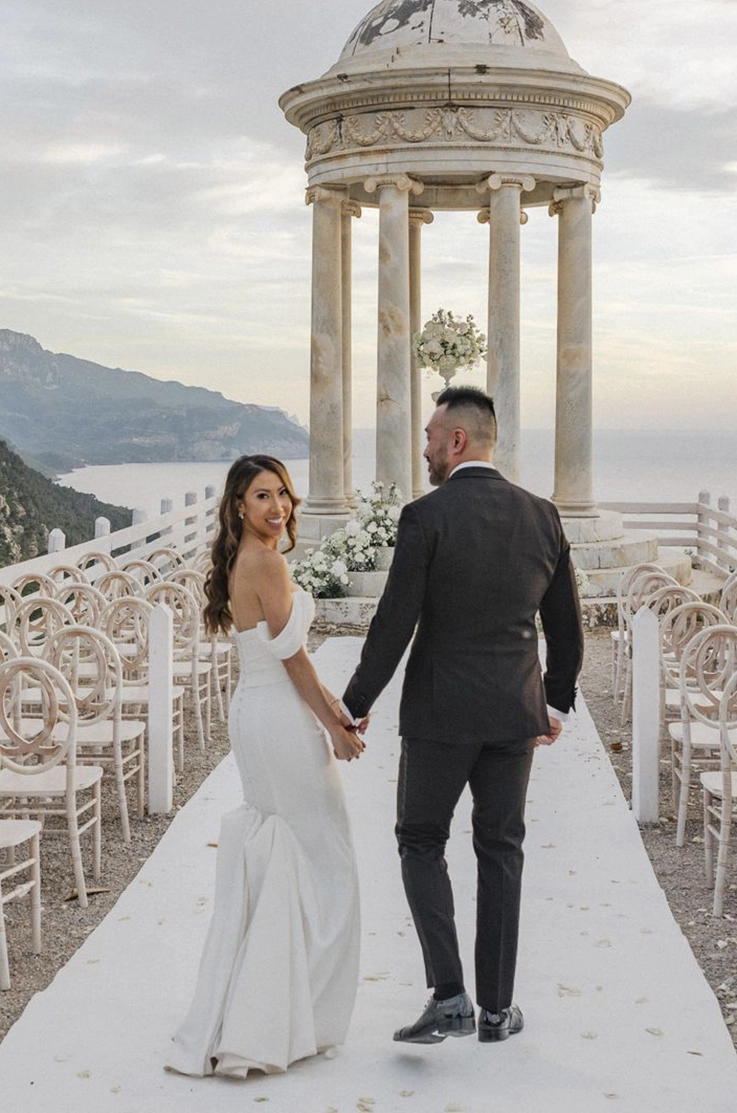 Bride looks back in a fitted crepe gown with draped sleeves, as she walks hand in hand with groom in the aisle by a cliffside gazebo. 