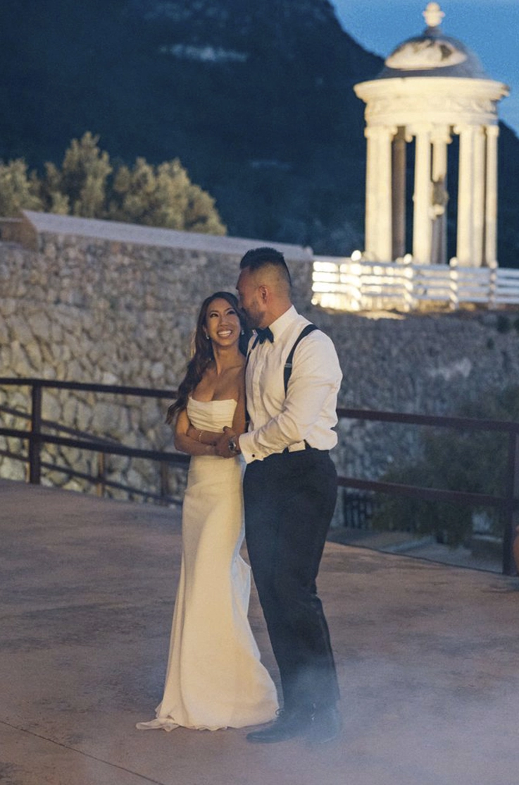 Groom embraces bride in a strapless fit and flare wedding dress with a draped crepe bodice, near mountaintop at dusk.